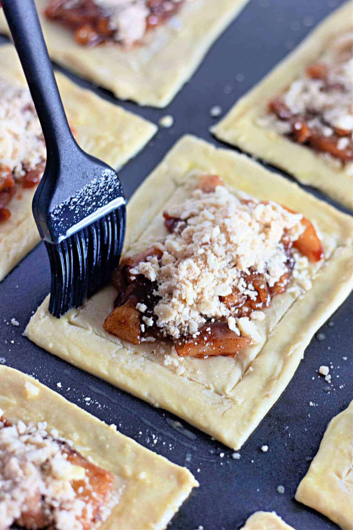 A black pastry brush brushing egg wash onto a rectangular unbaked apple danish topped with streusel.