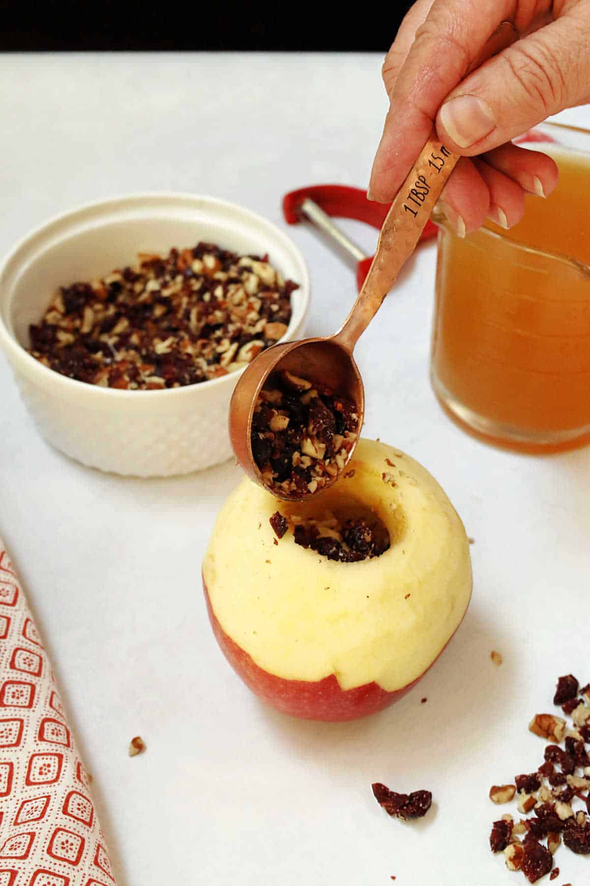 A hand fills a cored, peeled apple with cranberries and pecans using a tablespoon. A bowl of the mixture and a glass apple cider are in the background.