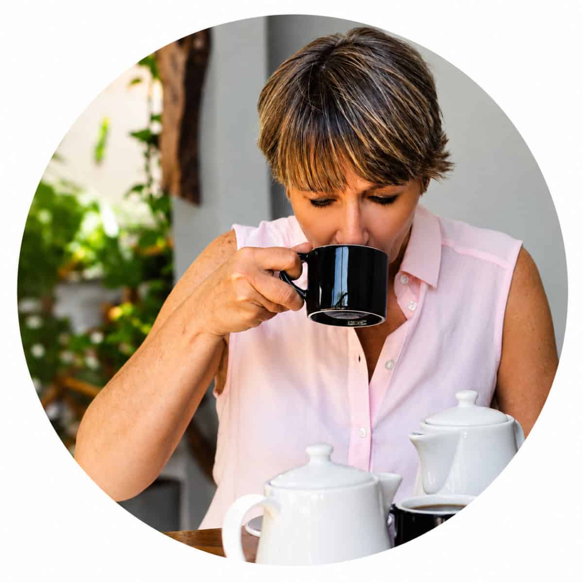 Jere in a sleeveless pink blouse drinks from a black mug at a table with teapots, with green plants in the background.