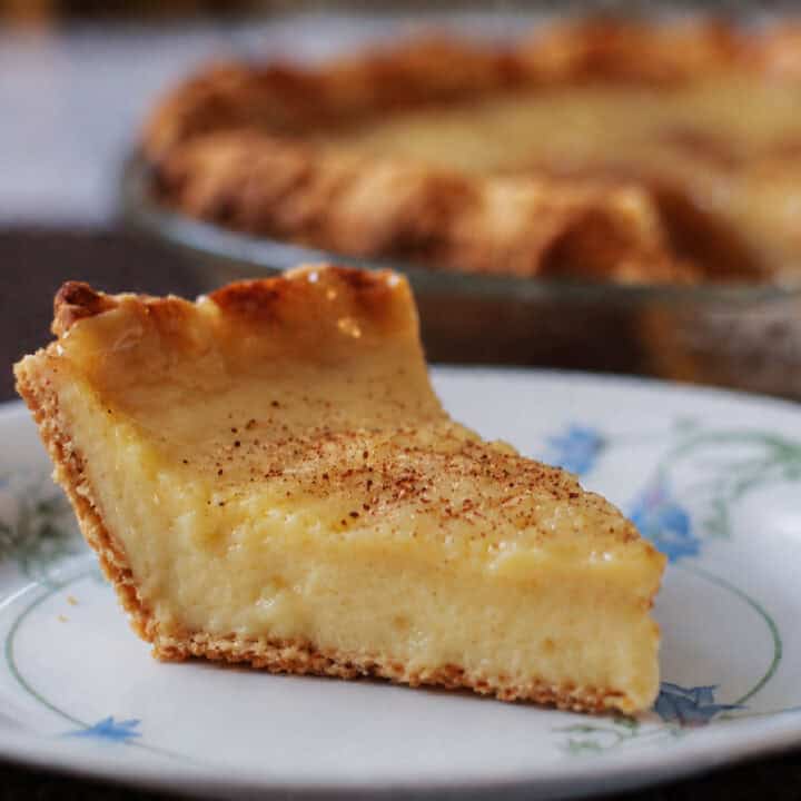 A slice of sugar cream pie with a golden crust sits on a decorative plate, with the rest of the pie visible in the background.