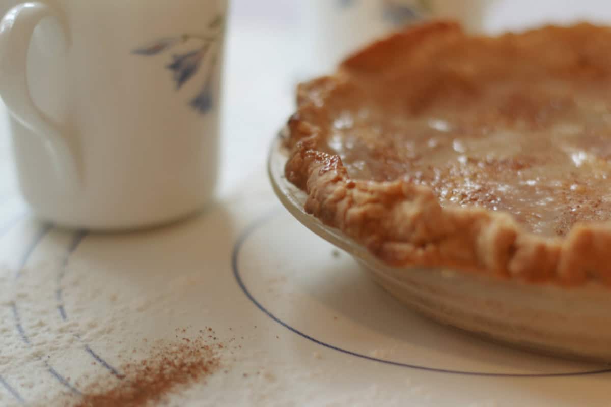 A close-up of a baked pie in a glass dish next to a white mug; some flour and cinnamon are sprinkled on the table.