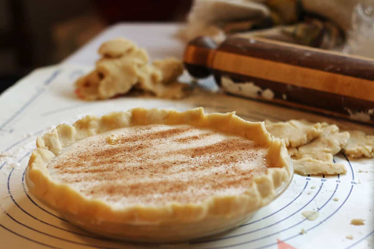 Unbaked sugar cream pie with crimped crust and sprinkled nutmeg sits on a pastry mat, surrounded by dough scraps and a rolling pin.