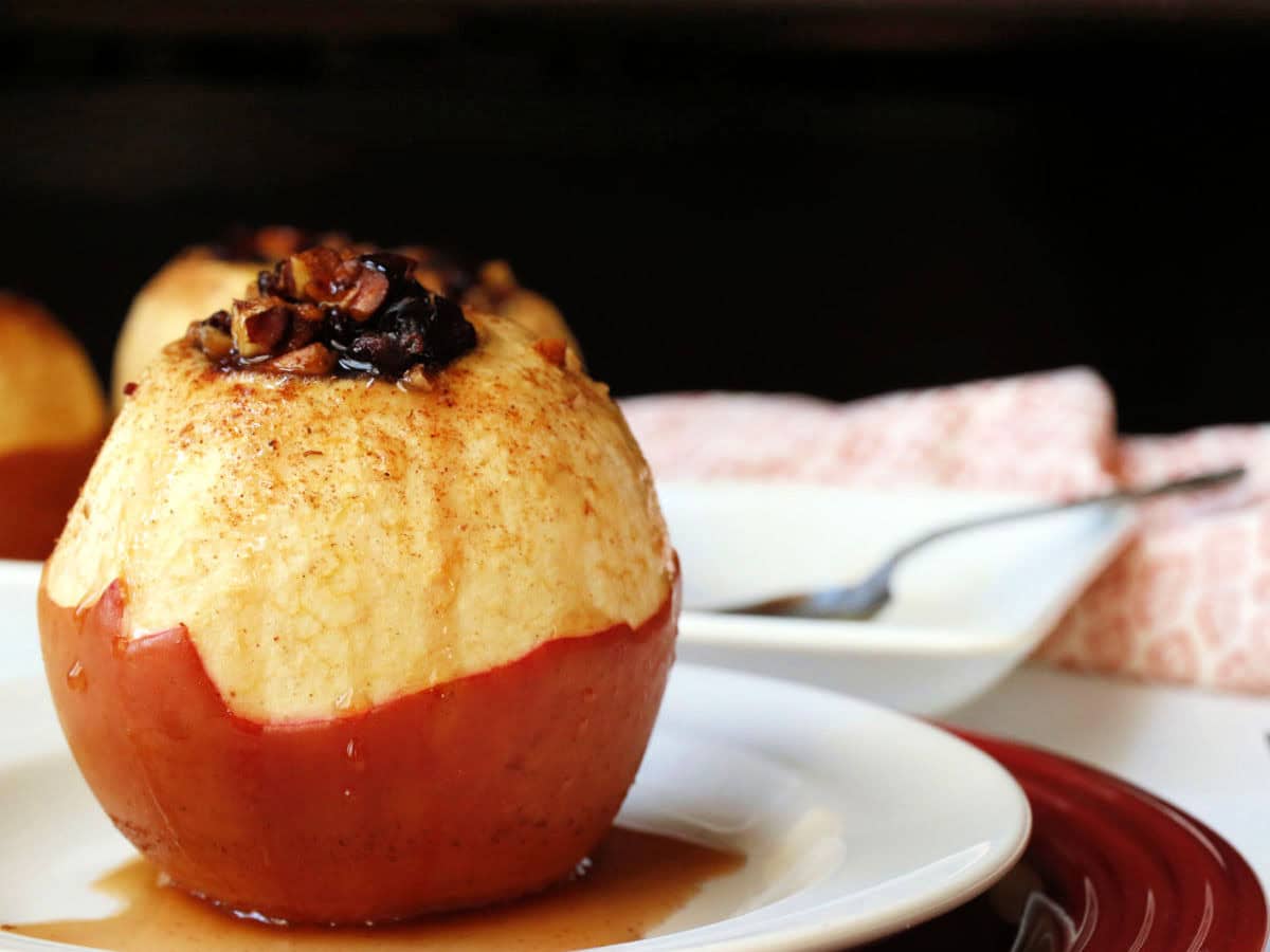 A baked apple with the top peeled, filled with cranberries and pecans, sits on a white plate with syrup, next to a fork and napkin.