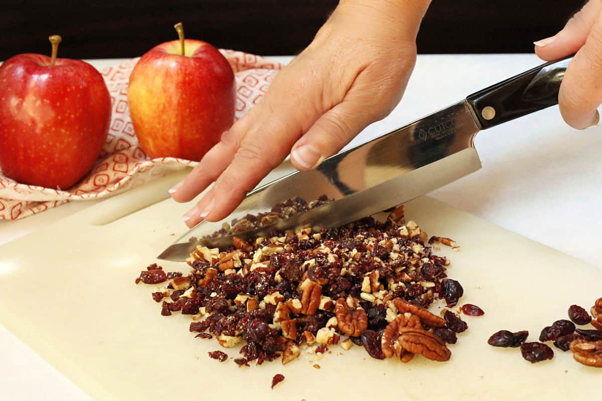 Hands chopping a mixture of dried cranberries and pecans on a cutting board, with two red apples and a patterned cloth in the background.