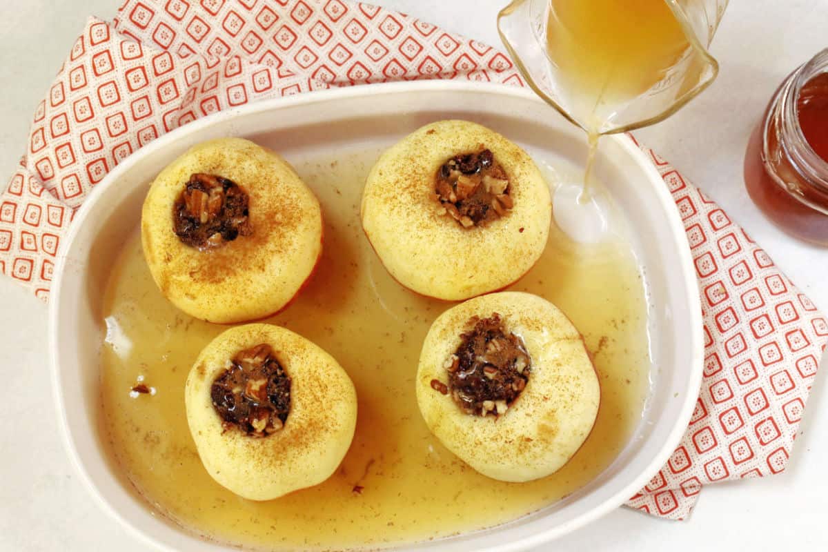 Four cored apples filled with nuts and dried fruit in a baking dish, with syrup being poured over them, on a red-patterned cloth.