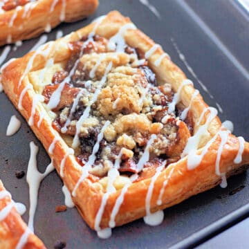 A rectangular apple danish pastry with a crumble topping and white icing drizzle on a baking sheet.