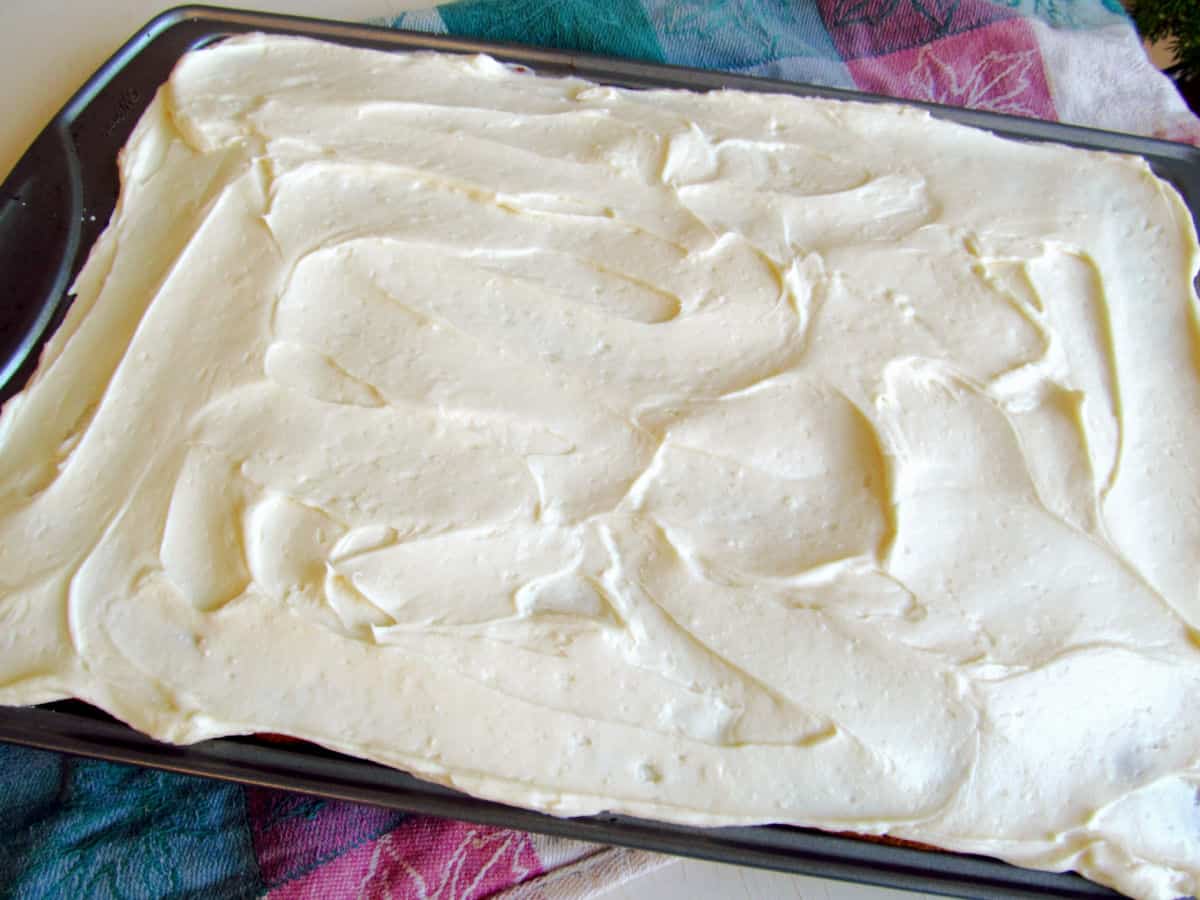 A pumpkin sheet cake with a layer of white, creamy frosting. The surface is spread unevenly with visible swirls and peaks. A colorful towel is partially visible underneath.