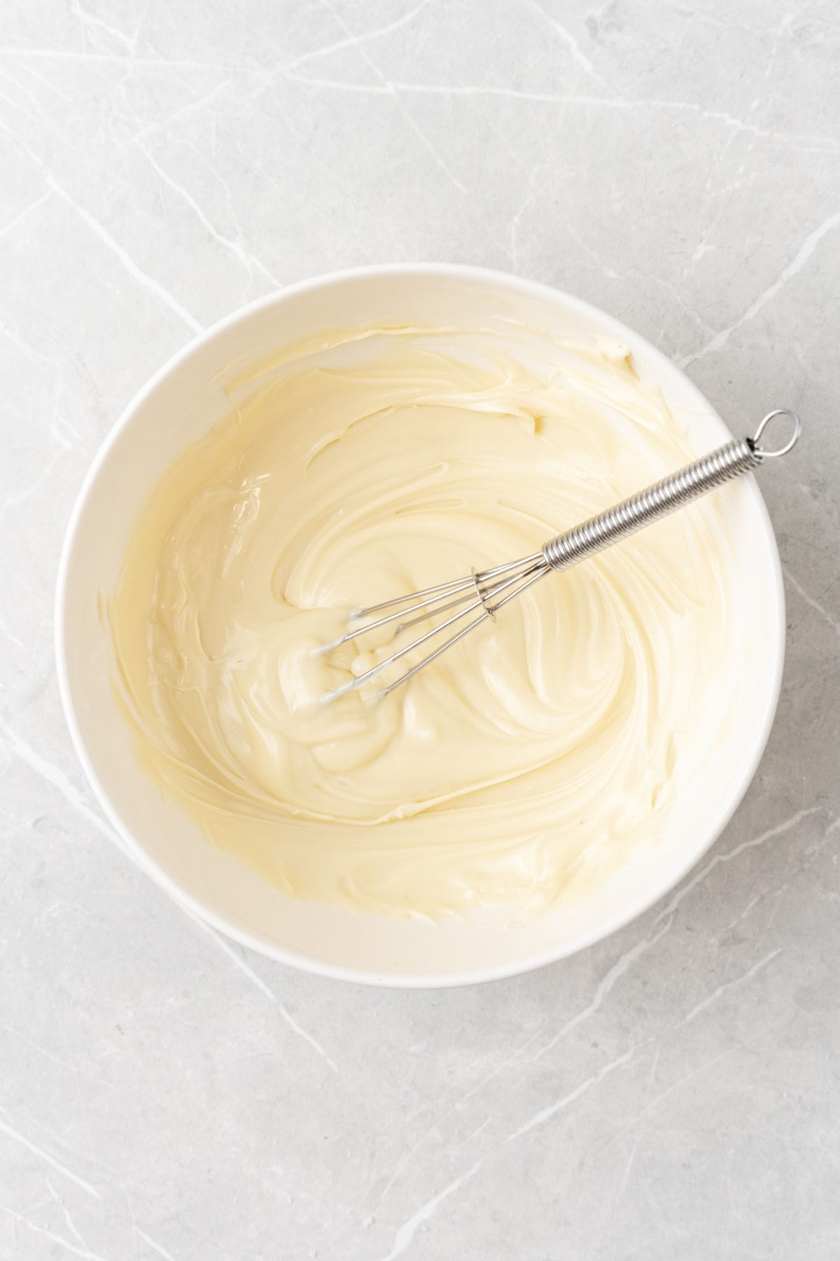 A white bowl filled with smooth melted white chocolate and a metal whisk resting inside, on a light gray marble surface.