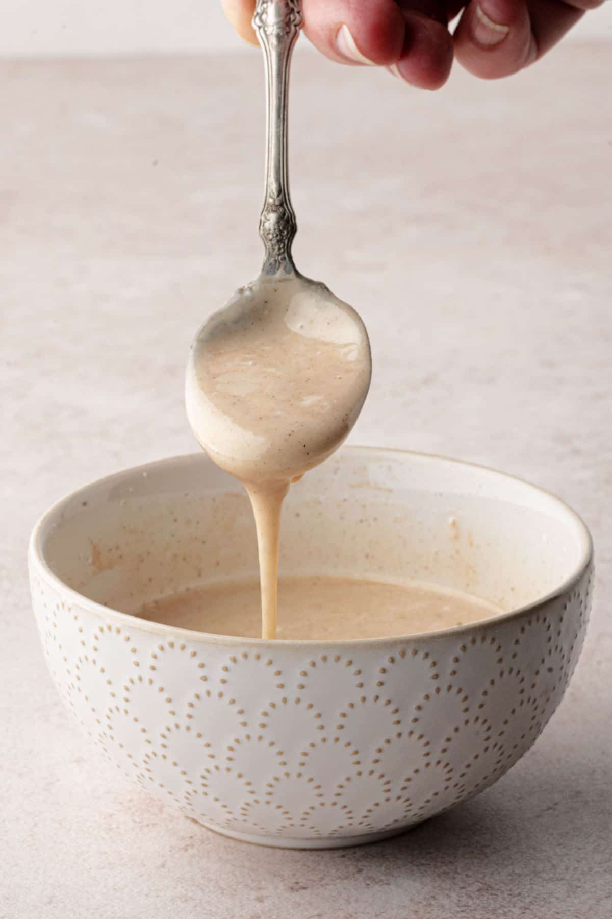 A hand holds a spoon drizzling light brown sauce into a patterned bowl.