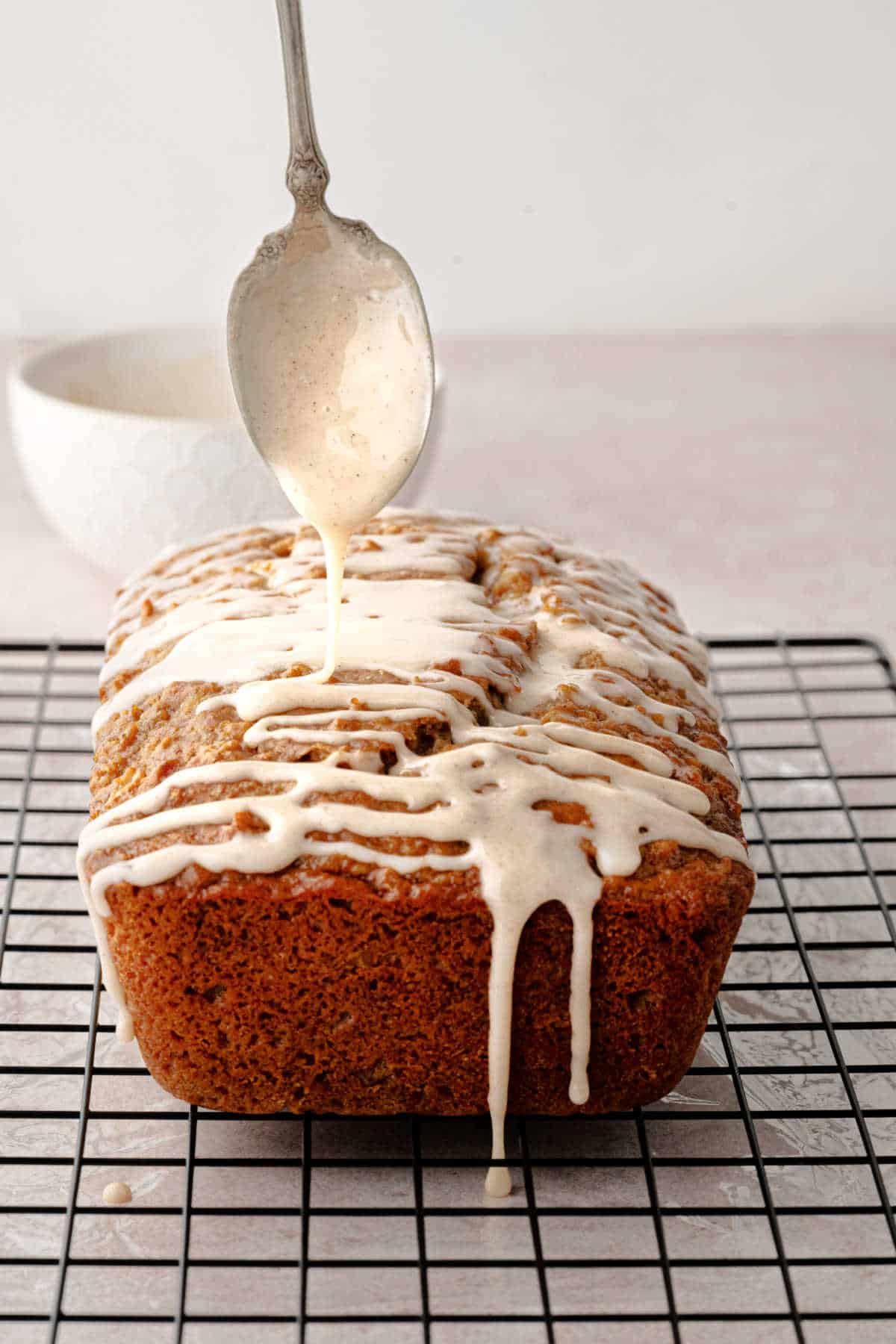 A metal spoon drizzles glaze over a loaf of baked bread sitting on a wire cooling rack.