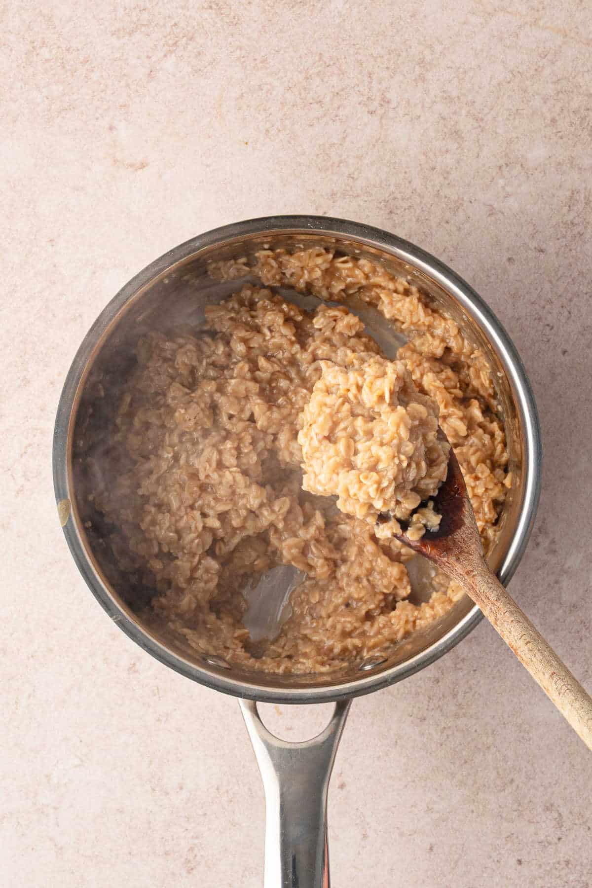 A wooden spoon lifts a scoop of cooked oatmeal from a metal saucepan.