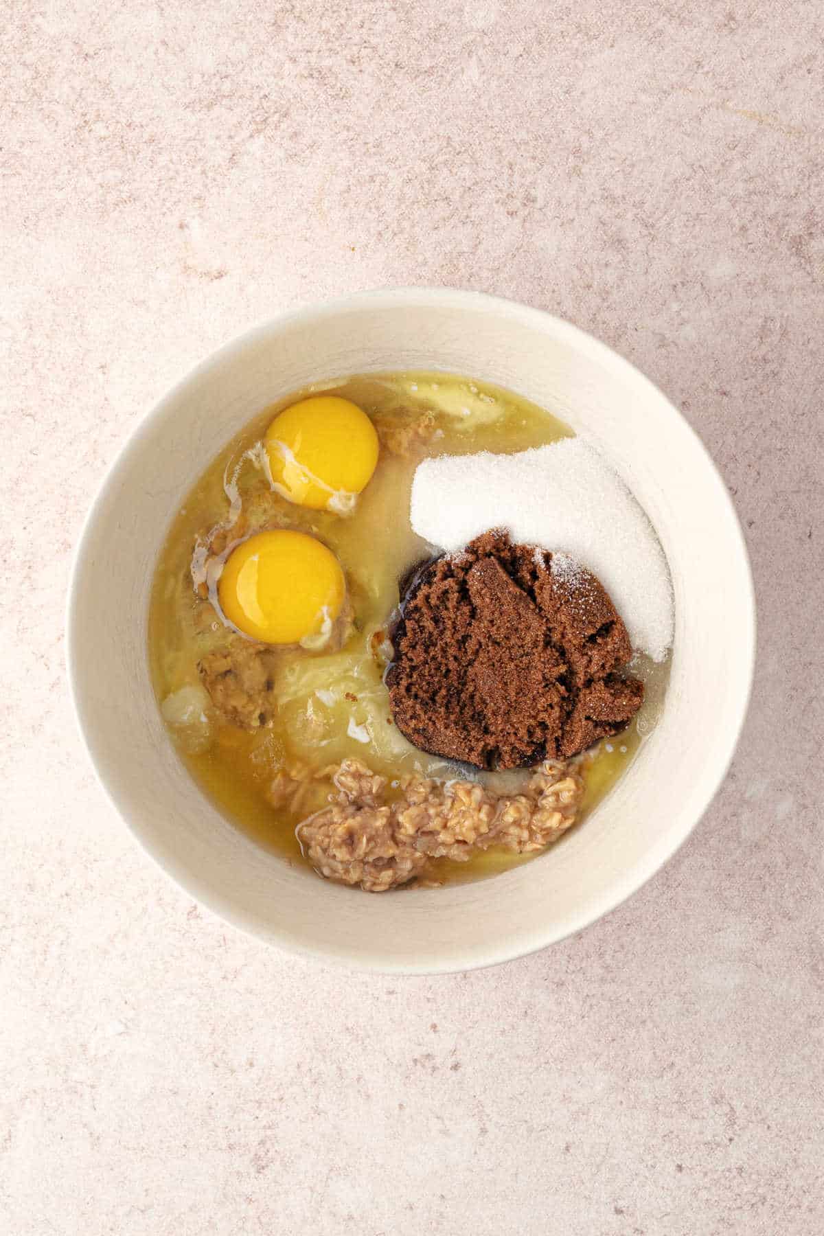 A bowl containing two raw eggs, white sugar, brown sugar, and rolled oats.