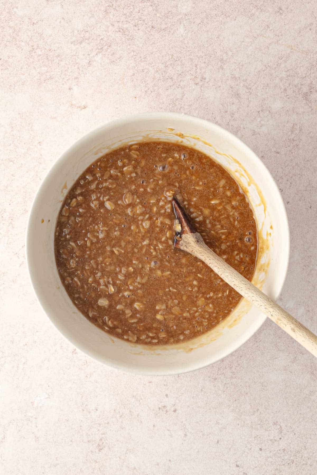A white bowl containing a brown, oatmeal-apple cider mixture being stirred by a wooden spoon.