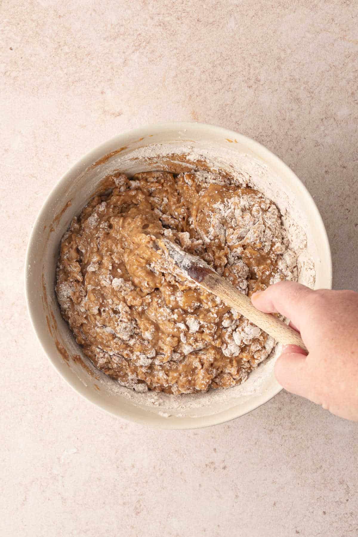 A hand uses a wooden spoon to stir flour into a bowl of wet cookie dough.