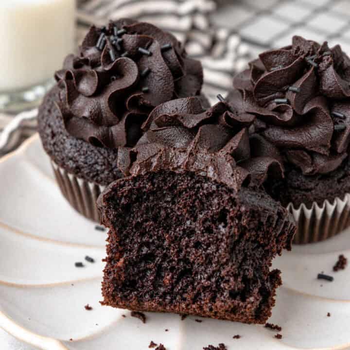Three chocolate black cocoa cupcakes with dark frosting and sprinkles on a white plate, one showing a bite taken out of it.