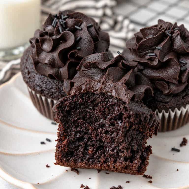 Three chocolate black cocoa cupcakes with dark frosting and sprinkles on a white plate, one showing a bite taken out of it.