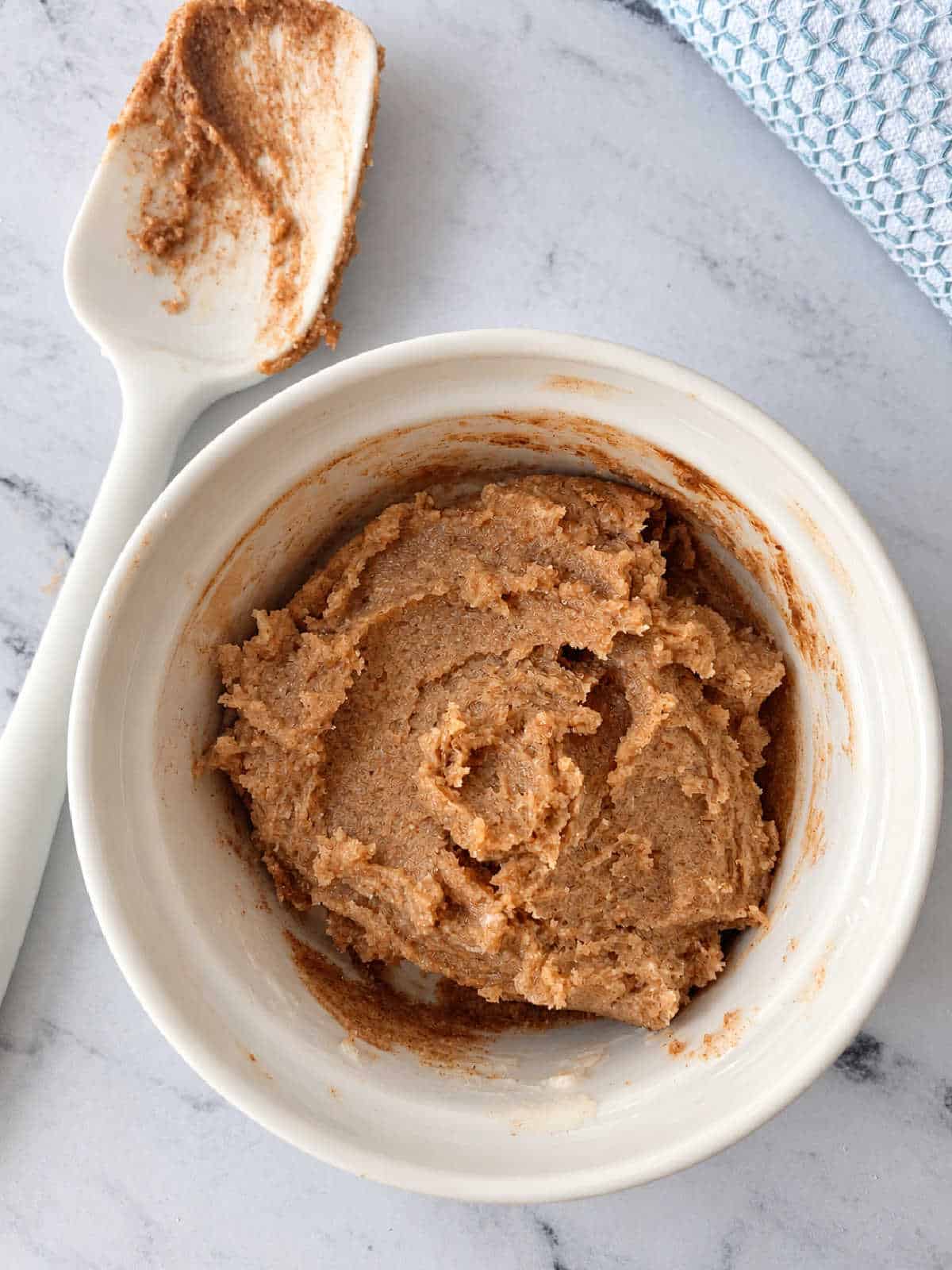 A small white bowl containing a thick, light-brown almond butter paste, with a white spatula resting nearby.