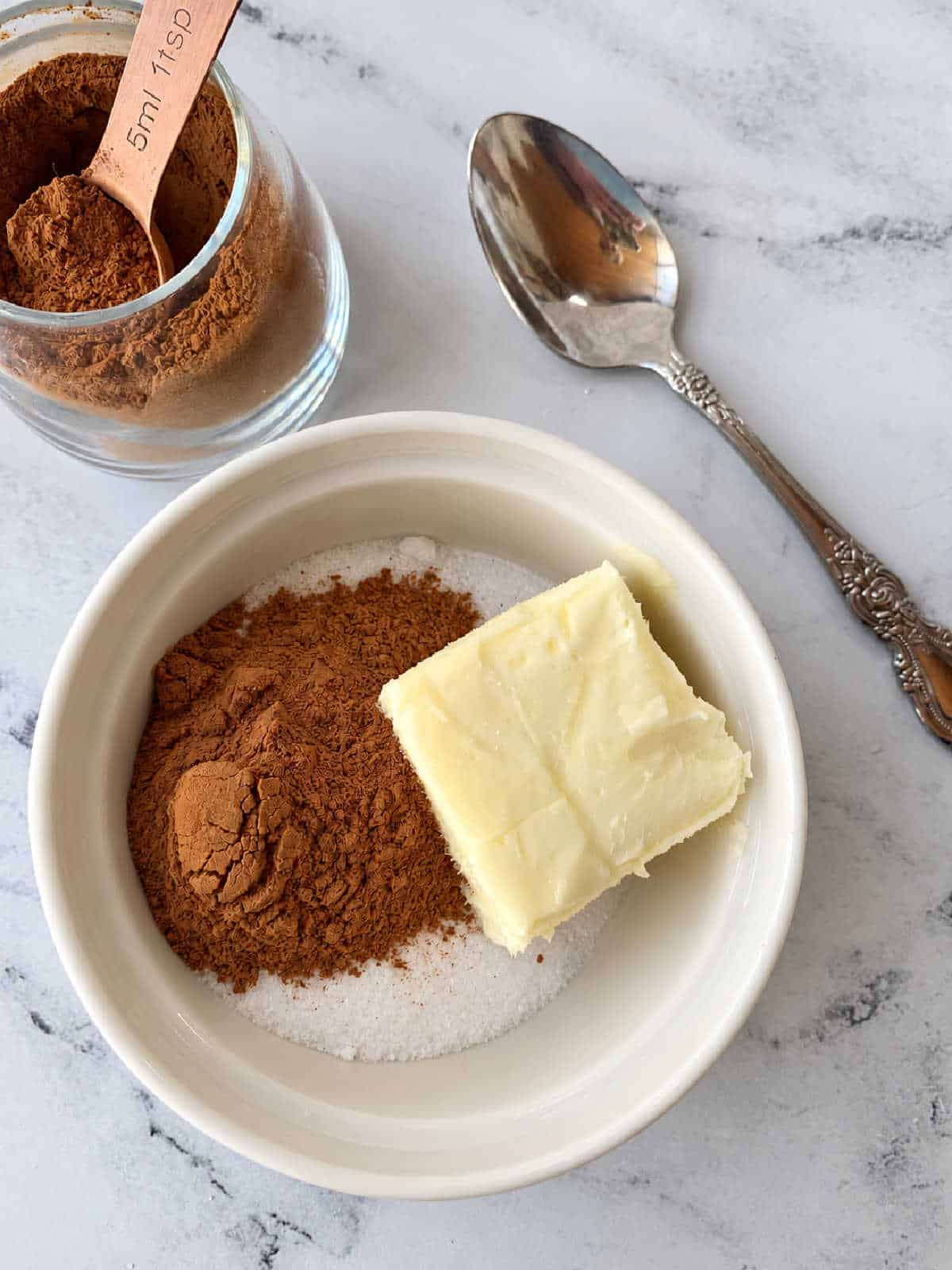 A white bowl containing cinnamon, sugar, and a square of butter, with a measuring spoon of cinnamon and a decorative metal spoon nearby.