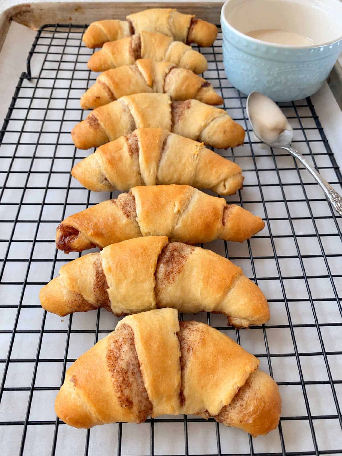 Seven crescent-shaped cinnamon rolls lined up on a wire cooling rack next to a bowl of icing and a spoon.