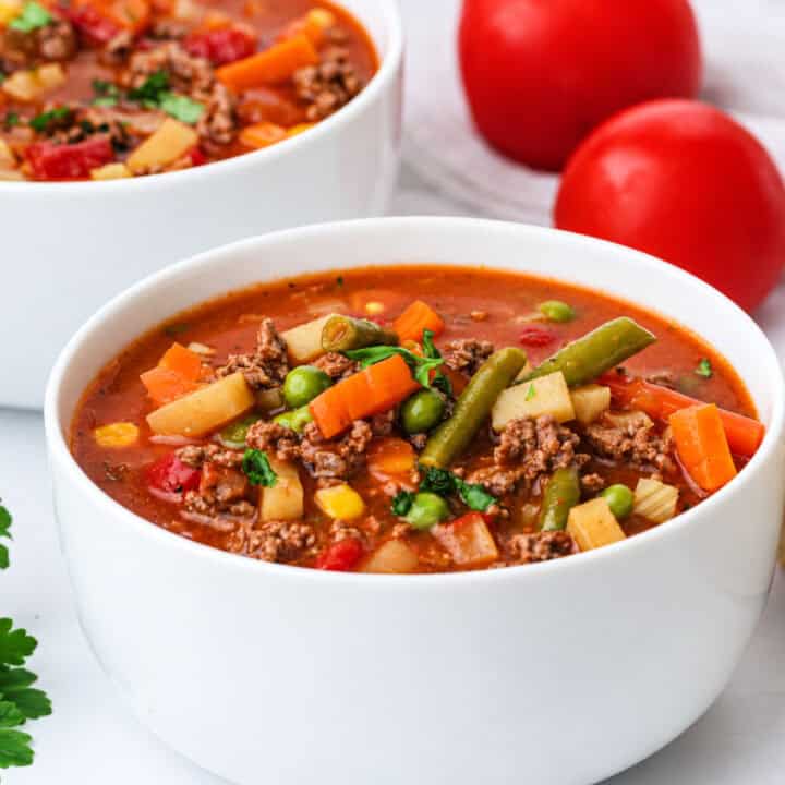Two bowls of vegetable beef soup garnished with herbs, with two whole tomatoes in the background.