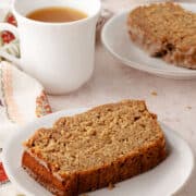 A slice of glazed banana bread on a small plate next to a mug of tea.