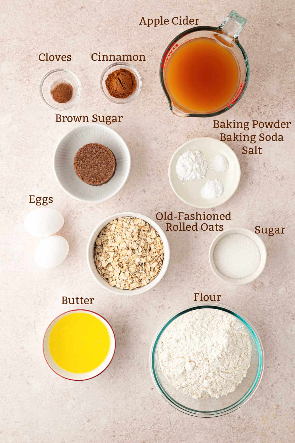 Ingredients for an apple cider oatmeal bread recipe arranged on a surface, labeled: apple cider, cloves, cinnamon, brown sugar, baking powder, baking soda, salt, eggs, oats, sugar, butter, and flour.